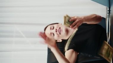 Dolarları saçan mutlu zengin kadın. Başarılı bayan patron ofise para saçıyor. Ofis masasında oturan bir kadın havaya para atıyor. Başarı ve zenginlik kavramı. Dikey video