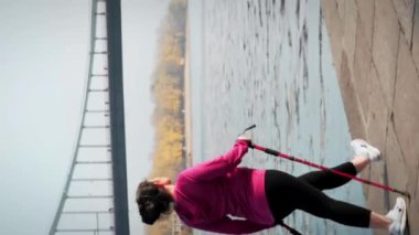 Siyah ve mor spor giyimli, İskandinav bastonlu tombul kadın eğitimini takip edin. Genç bir kadın sabahları dışarıda egzersiz yapıyor. Yoğun bir spor yapıyor. Dikey video