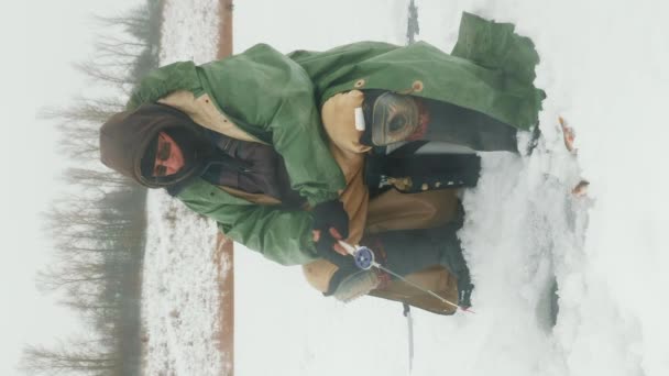 冬の極端な服を釣るロッドは 氷の穴で魚を捕まえる 凍った湖や川で冬の釣りをする漁師 バーティカルビデオ — ストック動画