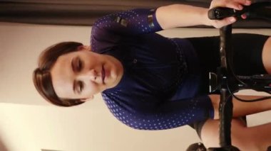 Kadın kapalı alanda bisiklet sürüyor, akıllı bisiklet çalıştırıyor, kardiyo egzersizleri yapıyor, içme suyu içiyor, kameraya bakıyor. Dikey video