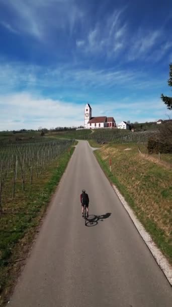 春にブドウ畑を通って道路自転車に乗る男 サイクリスト自転車は バックグラウンドに町役場を備えています バーティカルビデオ — ストック動画