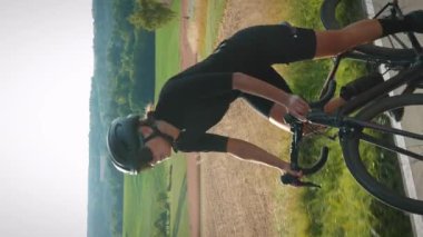 Gün batımında bisiklet süren şık formda bir bisikletçinin portresi. Dikey video. Bisiklet kasklı, gözlüklü dağlarda bisiklet süren, pedalları iten kendine güvenen bir kadın.