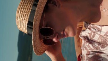 Güneş gözlüklü bir kadının portresi güneşli yaz gününde göl kenarındaki elektrikli turist teknesinde dinleniyor. Hasır şapkalı çekici bir kadın suyun üzerinde yüzüyor. Dikey video