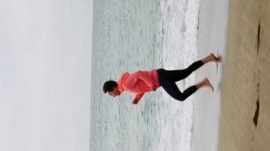 Turuncu ceketli genç bir kadın fırtınalı havada kumlu sahilde koşuyor. Büyük mavi dalgalar kuma çarpıyor. Kumda koşan bir kadın. Dikey video