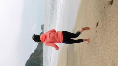 Kumsalda koşan kadın. Turuncu ceketli bir kadın deniz kıyısında koşuyor, kumlu sahilde dışarıda egzersiz yapıyor. Koşmak ve sağlıklı yaşam tarzı kavramı. Dikey video