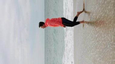 Turuncu ceketli ve taytlı bir kadın fırtınalı bir havada kumsalda yürüyor. Dikey video