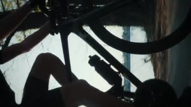 Erkek bacakları gün batımında bisiklet üzerinde pedal çeviriyor. Bisikletçi nehir kıyısı boyunca parkta bisiklet sürüyor. Spor şortlu erkek bacakları ve bisiklet ayakkabıları pedal çeviriyor. Dikey video