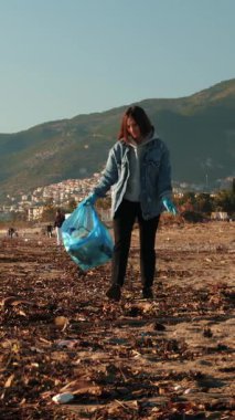Volunteer cleaning up beach from trash. Female collecting plastic bottles on beach. Woman picking up trash in trash bag. Global environment pollution. Vertical video