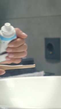 Close up of female hands in bathroom putting toothpaste on toothbrush, preparing to brush teeth. Woman puts toothpaste on brush, hold it over sink. Vertical video