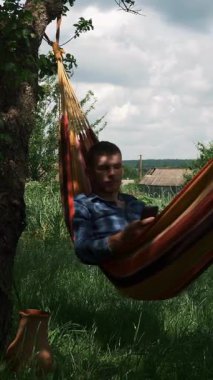 Man with smartphone in hands relaxing in hammock outdoors. Young male lying on hammock and using mobile phone. Camping. Summer vacation. Vertical video