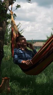 Handsome happy man talking by smartphone while lying in hammock. Male speaking on mobile phone and swinging in hammock. Relax in hammock. Summer vacation. Vertical video