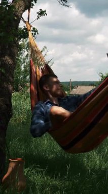 Man is relaxing in hammock, taking selfie photo on mobile phone. Man takes selfies on smartphone on hammock at green garden. Vertical video
