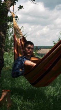 Handsome man relaxing in hammock and reading book at summer sunny day. Young male lying in hammock at green garden. Vertical video