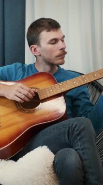 Gülümseyen kaygısız genç adam sıcak oturma odasında akustik gitar çalar. Erkek müzisyen evde sandalyede gitar çalıyor. Dikey video
