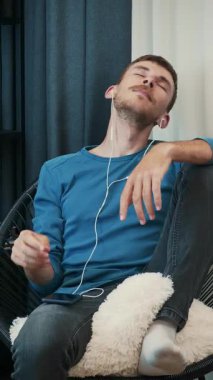 Tasasız, mutlu kulaklıklı, rahat koltukta oturmuş müzik dinleyen bir adam. Kulaklıklı bir erkek evde müzik dinliyor. Dikey video