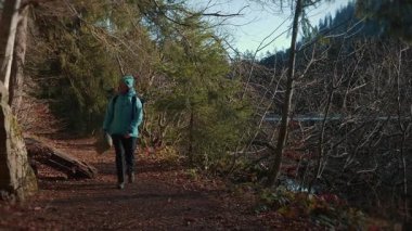 Smiling woman reading map while exploring nature, walking in sunny autumn forest along mountain lake. Female hiker with backpack hiking in nature park at sunny day, looking at map.