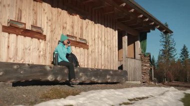 Woman sitting on bench near forest cabin, drinking tea and enjoying sunny day in forest. Female hiker exploring nature. Adventure travel. Active lifestyle concept