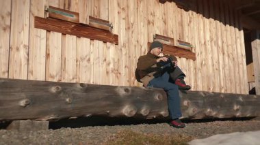 Male traveller pouring hot tea from insulated drink container, enjoying sunny autumn day outdoor. Man tourist sitting on bench near forest cabin and pouring tea from thermos