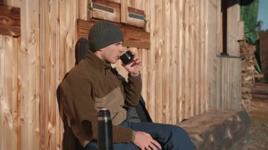 Portrait of young male tourist drinking hot tea from thermos while sitting on bench by forest cabin, enjoying sunny weather in fall forest. Man traveller making pause during hiking trip in mountains