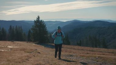 Woman with backpack climbing up mountain top at sunny fall day with mountain ranges on background. Female tourist in travel clothes is hiking outdoors. Active lifestyle concept