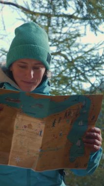 Portrait of female standing in forest and holding map in hands, looking for direction and enjoying nature. Young smiling woman exploring nature at sunny autumn day. Vertical video
