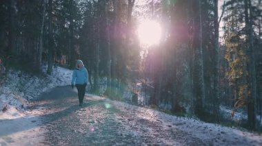 Mavi spor ceketli, şapkalı, sırt çantalı bir kadın güneşli kış gününde ormanda yürüyor. Ormanda yürüyüş yapan, doğayı keşfeden ve etrafa bakan bir kadın.