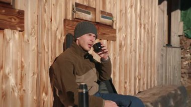 Turist spor kıyafetleri içinde orman kulübesinde çay içip güneşli bir günün tadını çıkaran bir adamın portresi. Yürüyüş ekipmanları içinde turist sıcak içecek içiyor, ufka bakıyor.