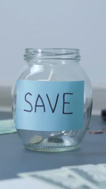 Coins falling into glass jar with word save. Transparent jar with save inscription on table among paper currency and coins. Vertical video
