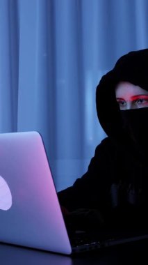 Woman face in black mask and hoodie looking at computer screen and typing on keyboard. Hacker breaks security protection on laptop. Vertical video