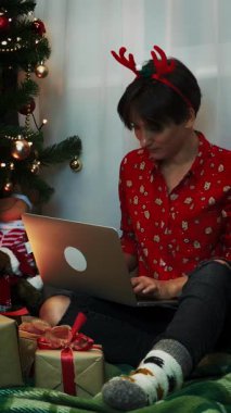 Brunette woman in Christmas party clothes is sitting near Christmas tree and working on computer at New Year night with lots of gift present boxes under trees. Vertical video