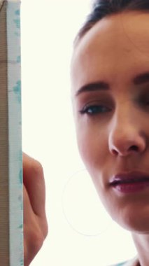 Portrait of young brunette woman with brush in hand painting picture on canvas in studio. Talented female working on abstract painting. Vertical video