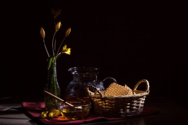 Hayat demlenmek için camdan bir çaydanlıktan, sarı nergis çiçeklerinden, kurabiyeli bir hasır sepetten, koyu arkaplanda bal olan şeffaf bir bardaktan oluşur.