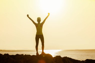 Deniz kenarındaki kayalıklarda gün doğumuna kollarını açan kadın.