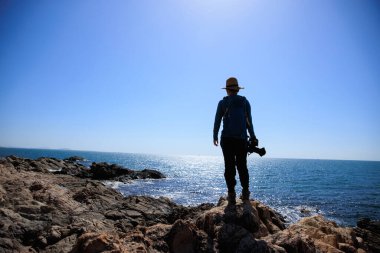 Kadın fotoğrafçı, deniz kenarındaki kayalıklarda güneşin doğuşunu seyrediyor.