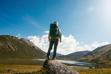 Sırt çantalı bir kadın yüksek irtifada dağ tepesinde yürüyüş yapıyor. Uzakta bir göl var.