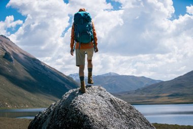 Sırt çantalı bir kadın yüksek irtifada dağ tepesinde yürüyüş yapıyor. Uzakta bir göl var.