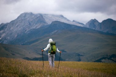Sırt çantalı bir kadın arka planda karlı dağlarla yüksek irtifada yürüyüş yapıyor.