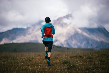 Fitness kadın patika koşucusu arka planda karlı dağlarla çayırlarda koşuyor.