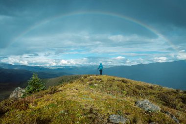 Sırt çantalı kadın yağmurdan sonra yüksek irtifada dağ tepesinde yürüyüş yapıyor, güzel bir gökkuşağıyla karşı karşıya.