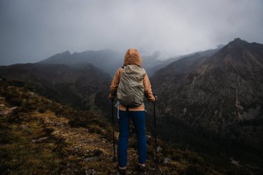 Sırt çantalı bir kadın yüksek irtifada dağ yürüyüşü yapıyor. Yağmurda karla kaplı dağa bakıyor.