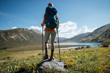 Sırt çantalı bir kadın yüksek irtifada dağ tepesinde yürüyüş yapıyor. Uzakta bir göl var.