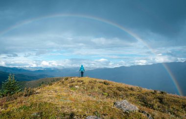 Sırt çantalı kadın yağmurdan sonra yüksek irtifada dağ tepesinde yürüyüş yapıyor, güzel bir gökkuşağıyla karşı karşıya.