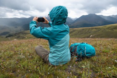 Kadın yürüyüşçü, yüksek irtifada dağların tepesinde fotoğraf çekmek için akıllı telefon kullanır.