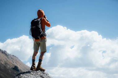 Kadın fotoğrafçı yüksek irtifada dağın tepesinde fotoğraf çekiyor. Uzakta bir göl var.