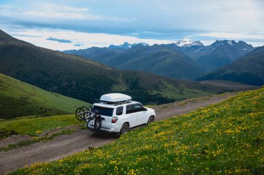 Offroad arabası mavi gökyüzünün altında çiçek açan çayır dağında bisiklet taşıyor.