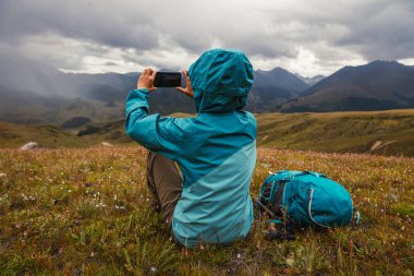 Kadın yürüyüşçü, yüksek irtifada dağların tepesinde fotoğraf çekmek için akıllı telefon kullanır.