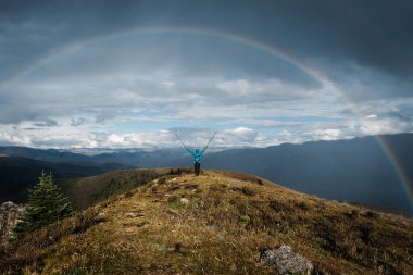 Başarılı bir sırt çantalı kadın yağmurdan sonra yüksek irtifada dağ tepesinde yürüyüş yaparken güzel bir gökkuşağıyla karşılaşıyor.