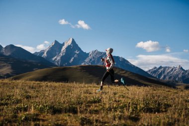 Fitness kadın patika koşucusu arka planda karlı dağlarla çayırlarda koşuyor.