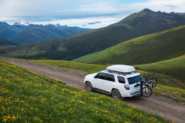 Offroad arabası mavi gökyüzünün altında çiçek açan çayır dağında bisiklet taşıyor.