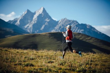 Fitness kadın patika koşucusu arka planda karlı dağlarla çayırlarda koşuyor.